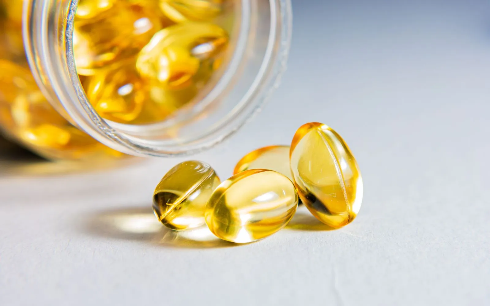 Clear golden softgel capsules spilling from a tipped-over glass jar onto a pale grey surface