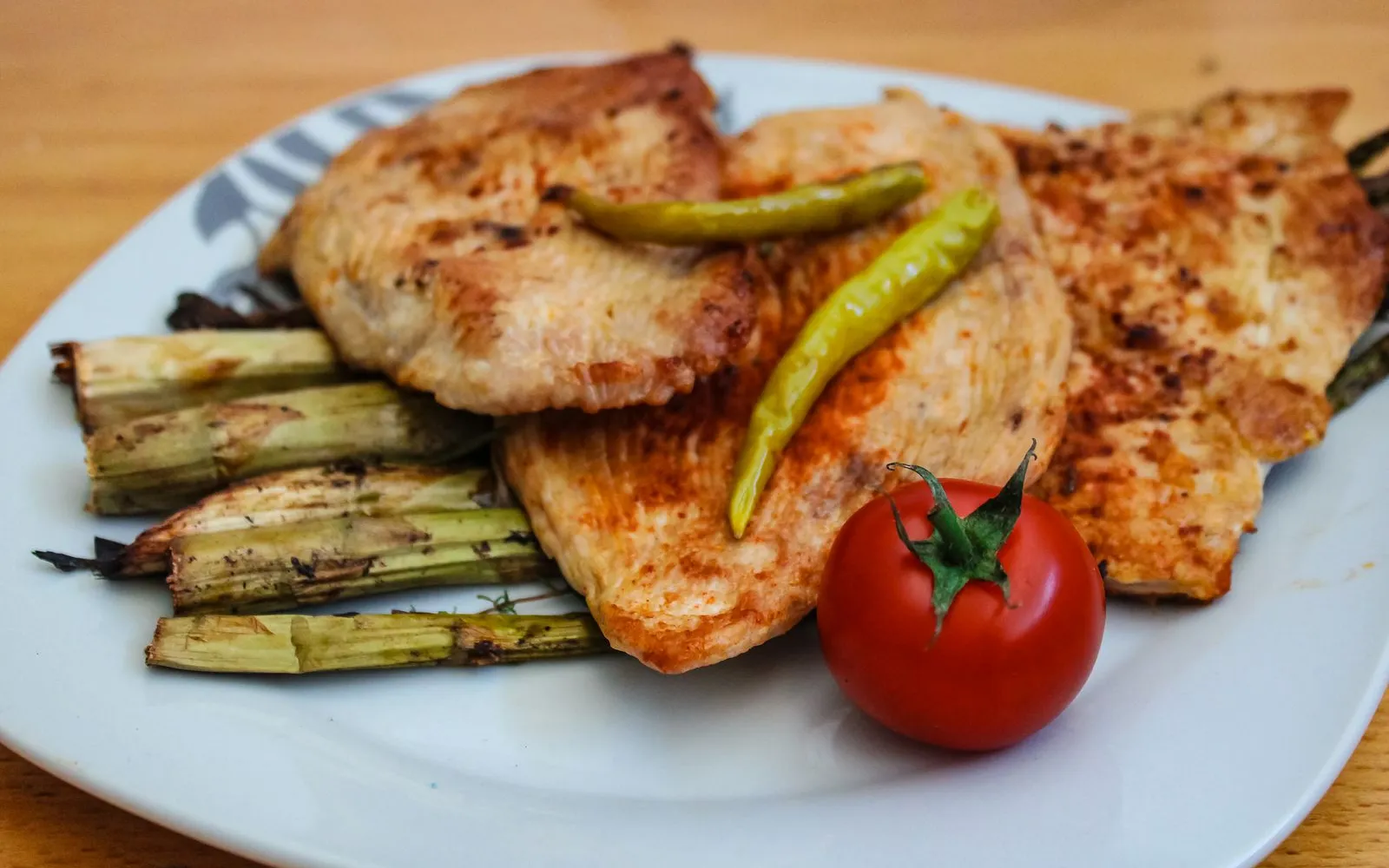 Grilled chicken breasts with roasted green vegetables, two green chillies and a cherry tomato on a white plate