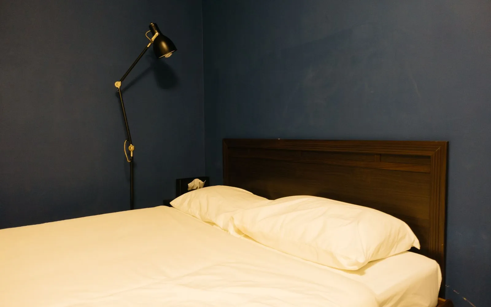 A made bed with white sheets and pillows against a dark blue wall, with a black floor lamp beside it