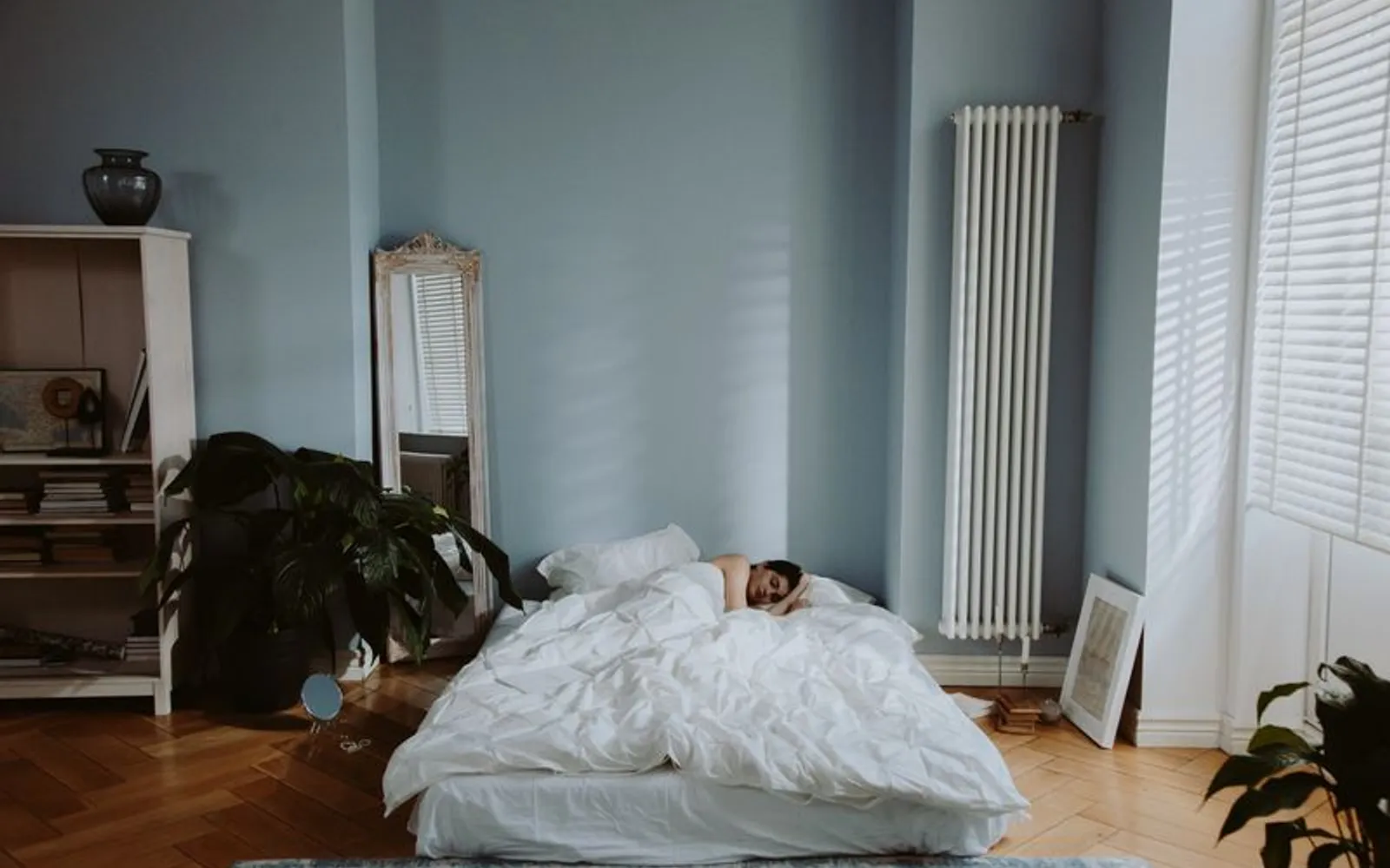 A person asleep under a white duvet on a low bed in a calm, pale blue bedroom