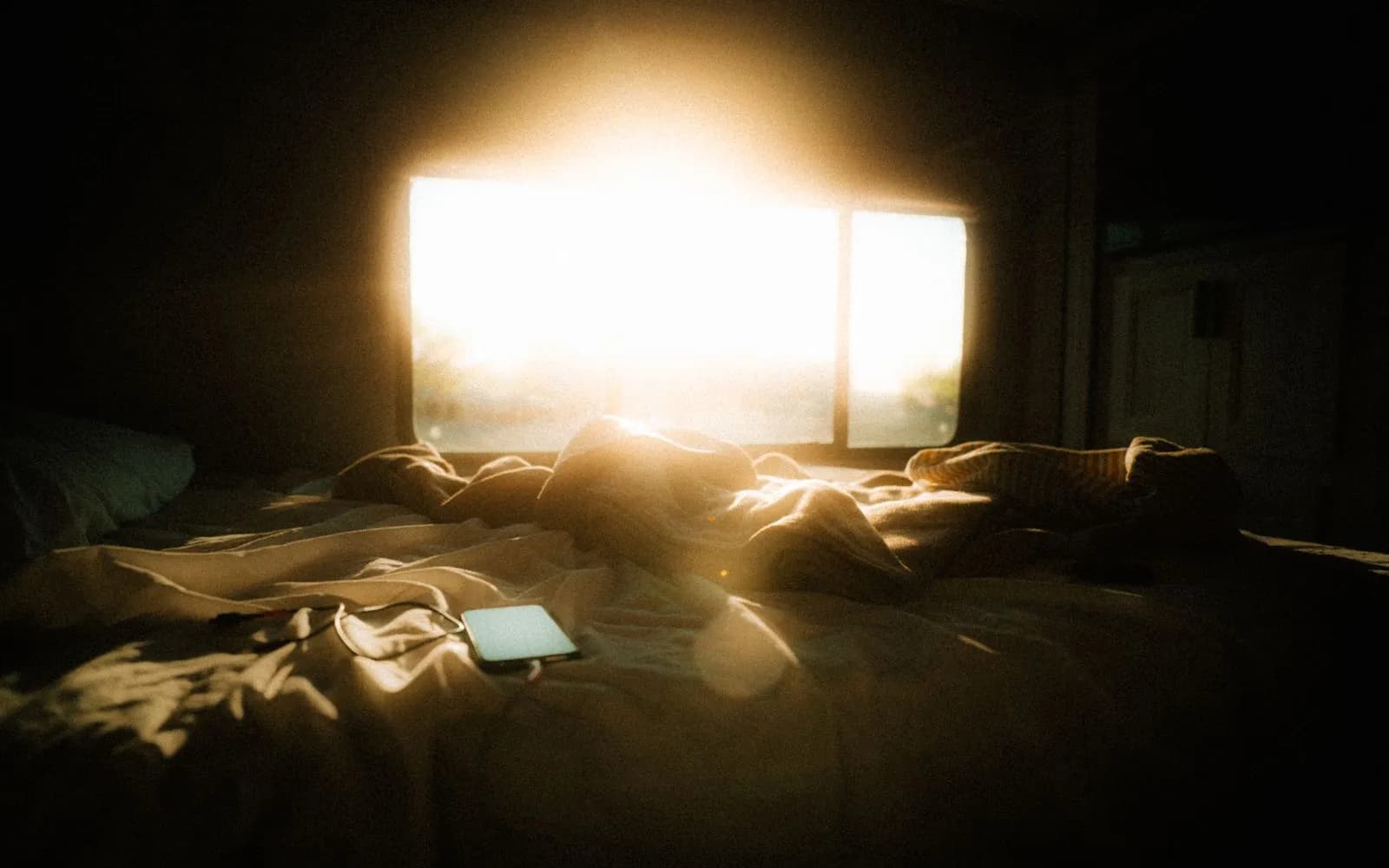 An unmade bed with rumpled covers and a phone lying on it, lit by bright morning sun through a window