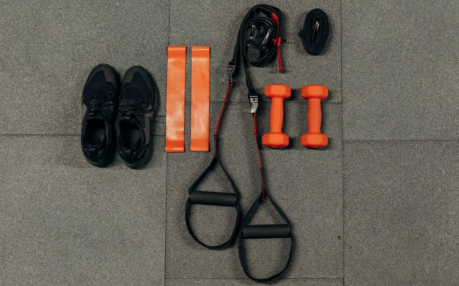 Two small orange dumbbells, two orange loop resistance bands, suspension straps and a pair of black trainers laid out on a grey rubber floor