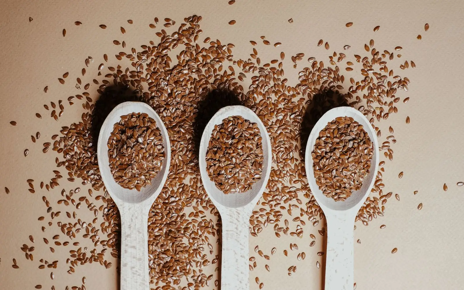 Three wooden spoons filled with brown flaxseeds, with loose seeds scattered on a beige surface