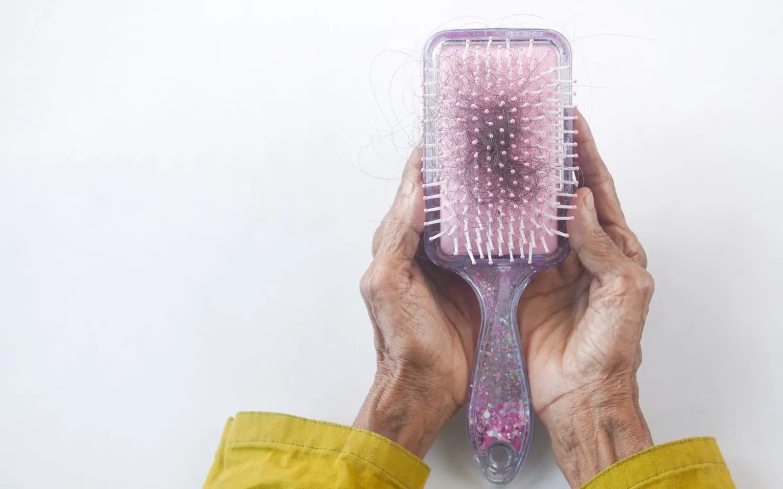 Two older hands holding a pink hairbrush full of loose dark hair against a white background