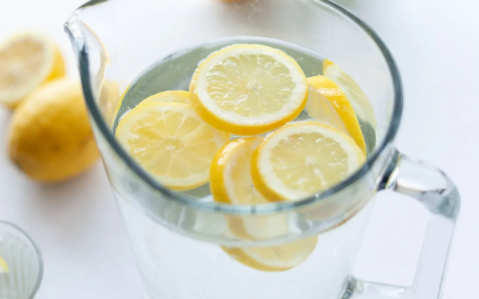 A glass jug of water with lemon slices, two empty glasses, whole lemons and a sprig of mint on a white table