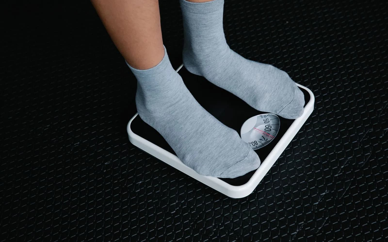 Feet in grey socks standing on a white dial bathroom scale on a dark rubber floor