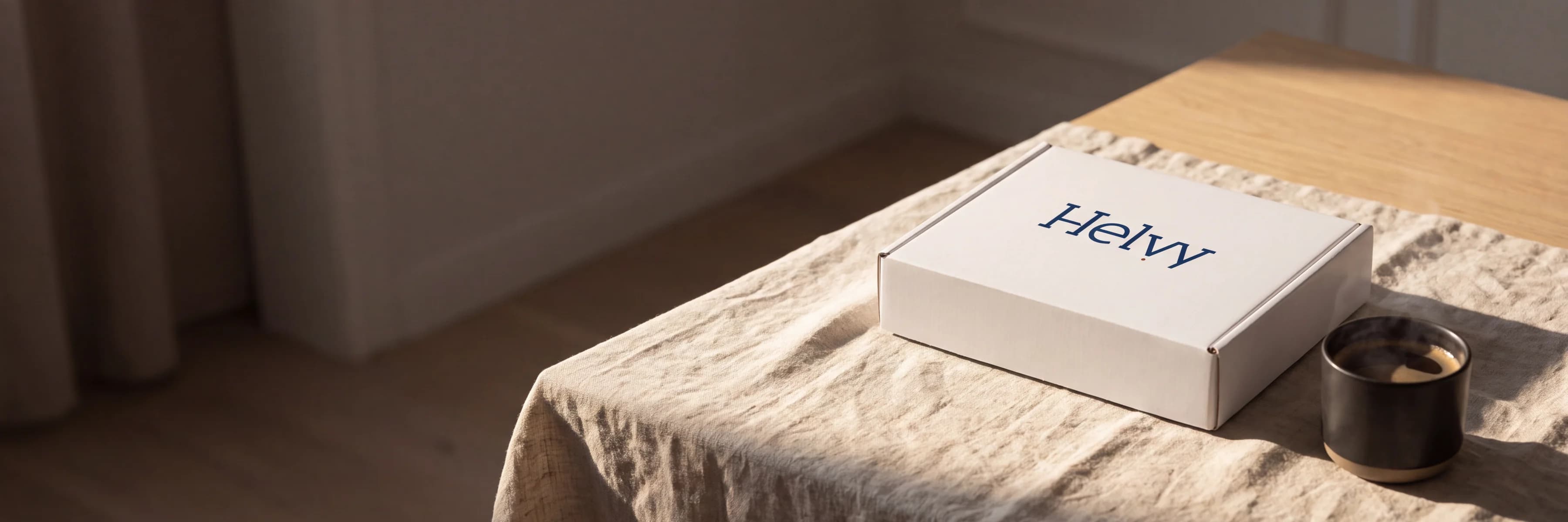 The white Helvy box and a dark stoneware cup on a linen cloth in raking morning light