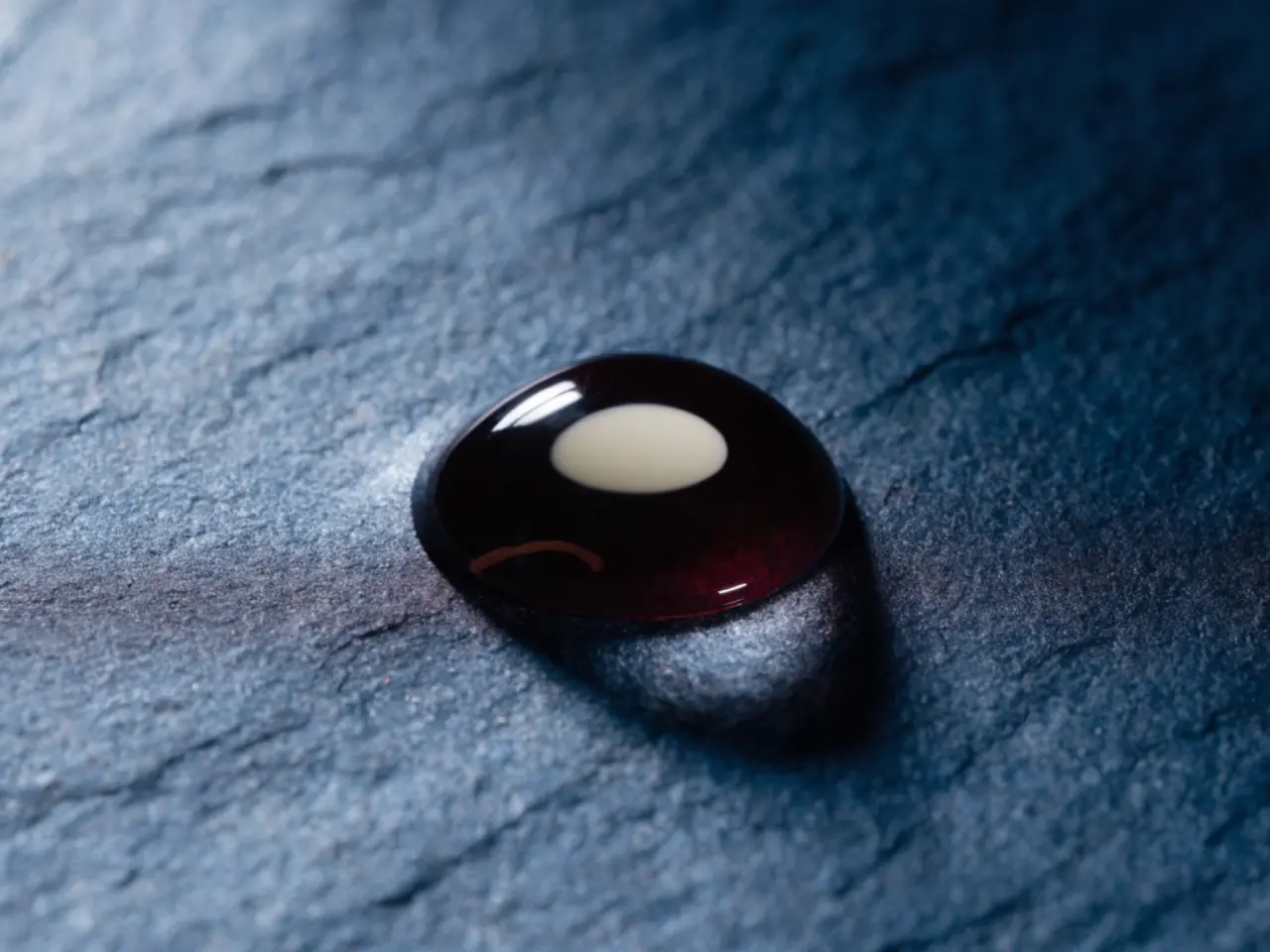 A single drop of blood resting on dark slate, lit from the side