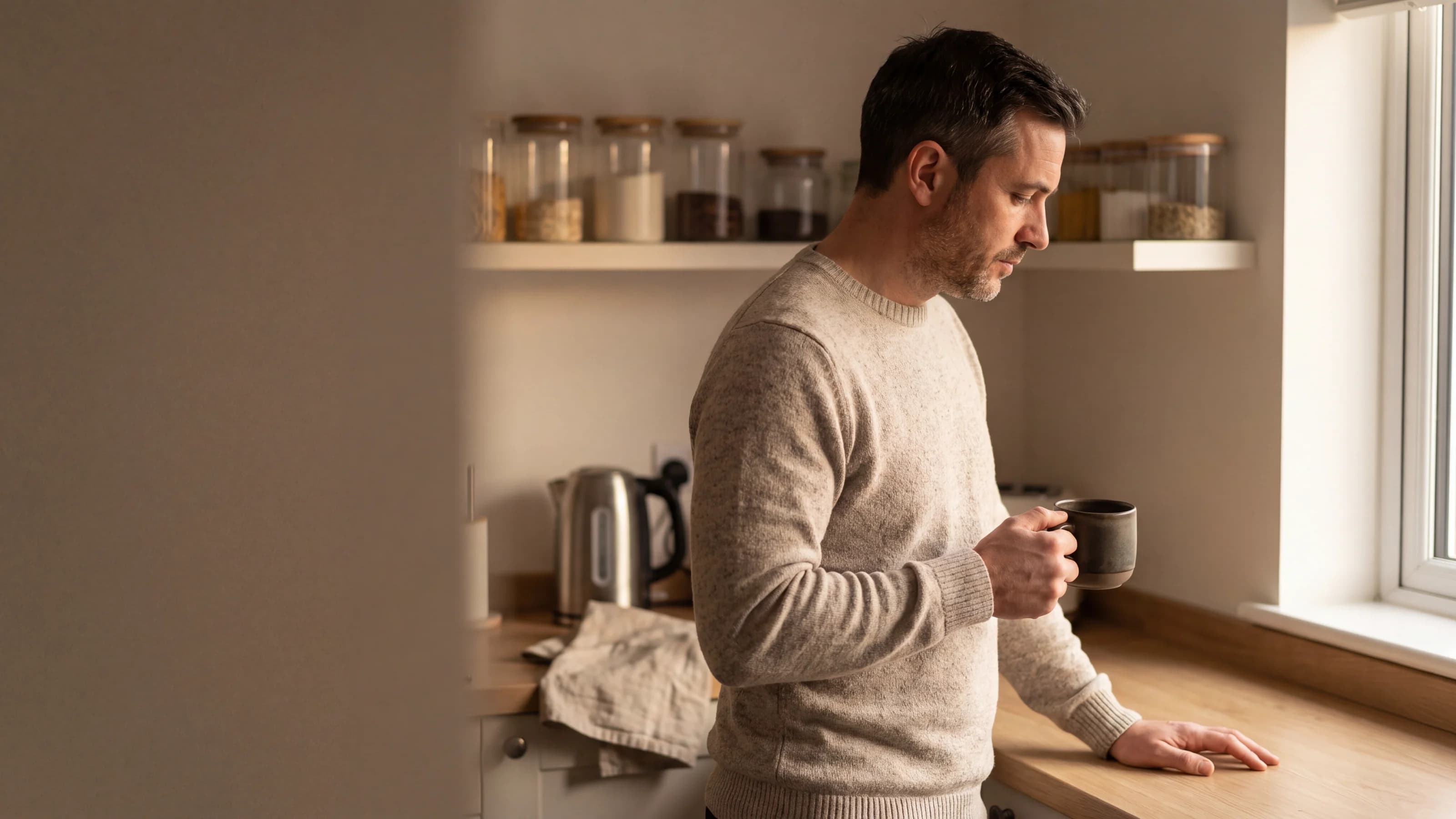 A man stands at a kitchen window holding a mug, looking out, morning light across the worktop