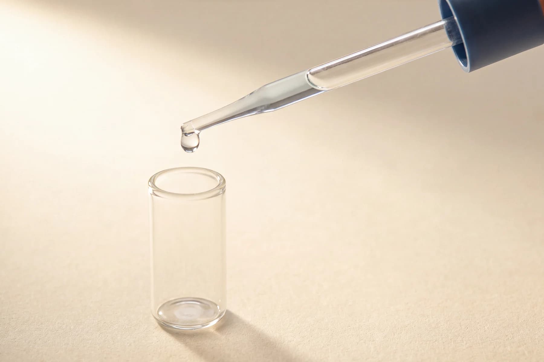 A glass pipette releasing a single drop into an open sample tube on a warm ivory surface