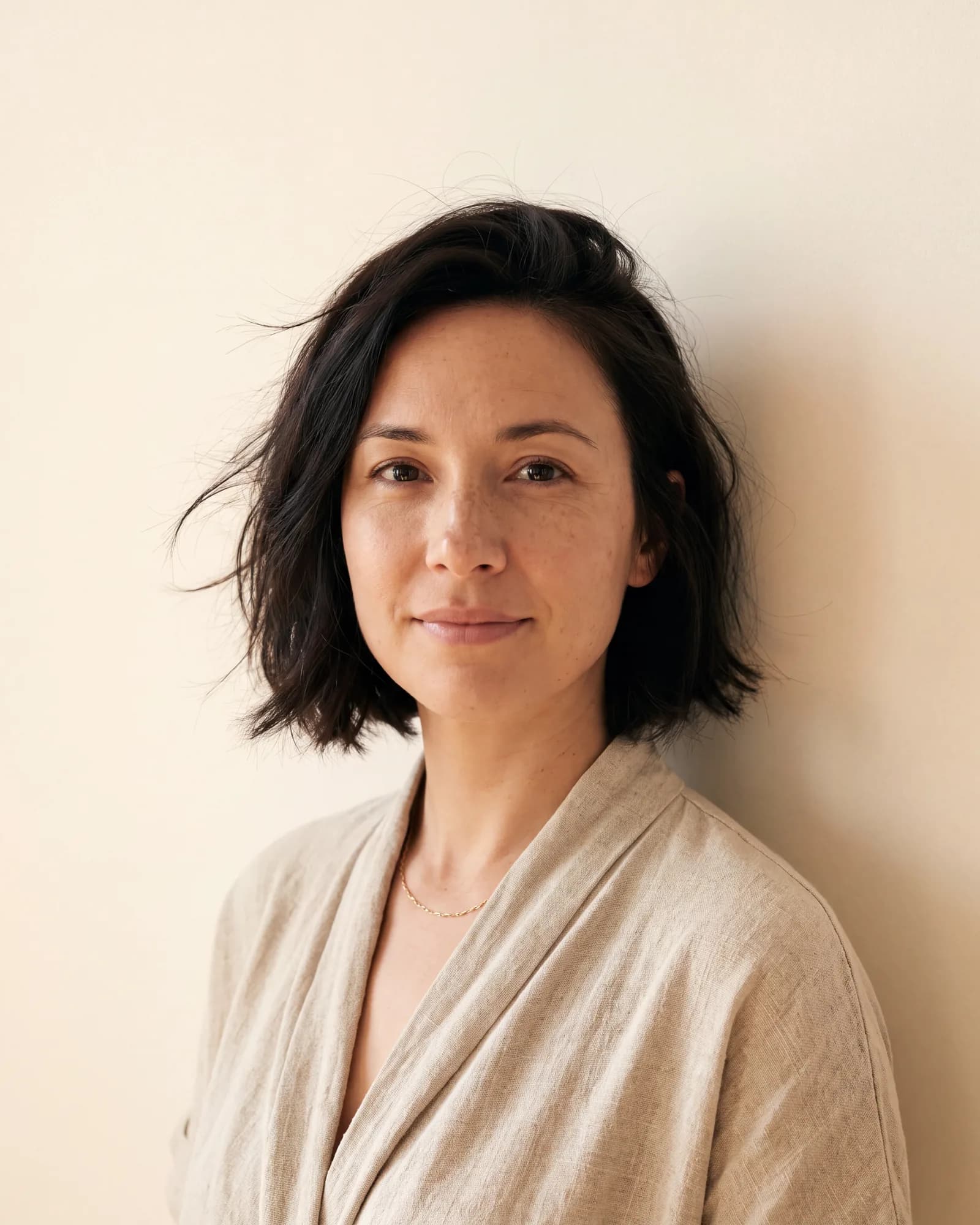 A woman in her thirties in an oatmeal knit stands against a warm ivory wall, half smiling to camera