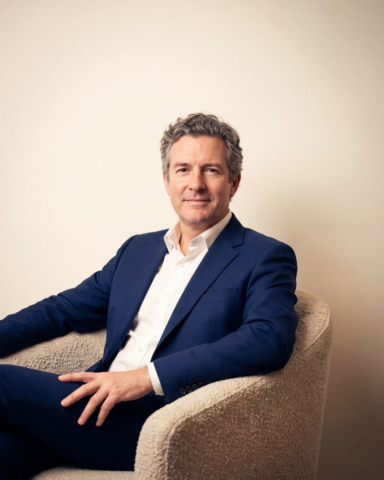 A man in his fifties with greying hair sits in a low bouclé armchair in an empty room, looking to camera